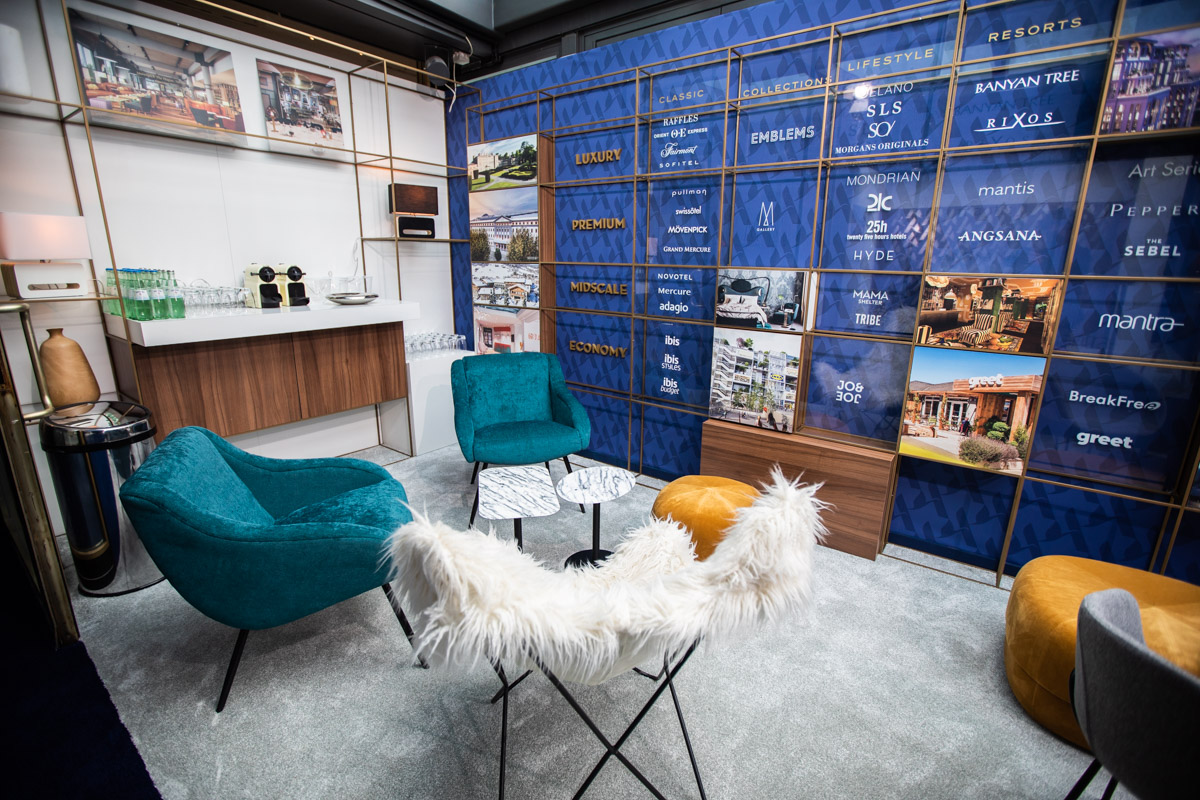 Accor at IHIF Berlin 2021 — Hospitality space Hospitality lounge with teal chairs, white seating and a blue hotel-brand display wall at IHIF Berlin 2021